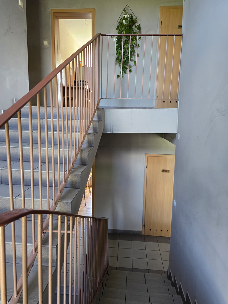 Klatka schodowa z metalową balustradą, drzwiami do pokoi i zieloną rośliną ozdobną na ścianie – Noclegi Gorzów w Baczynie